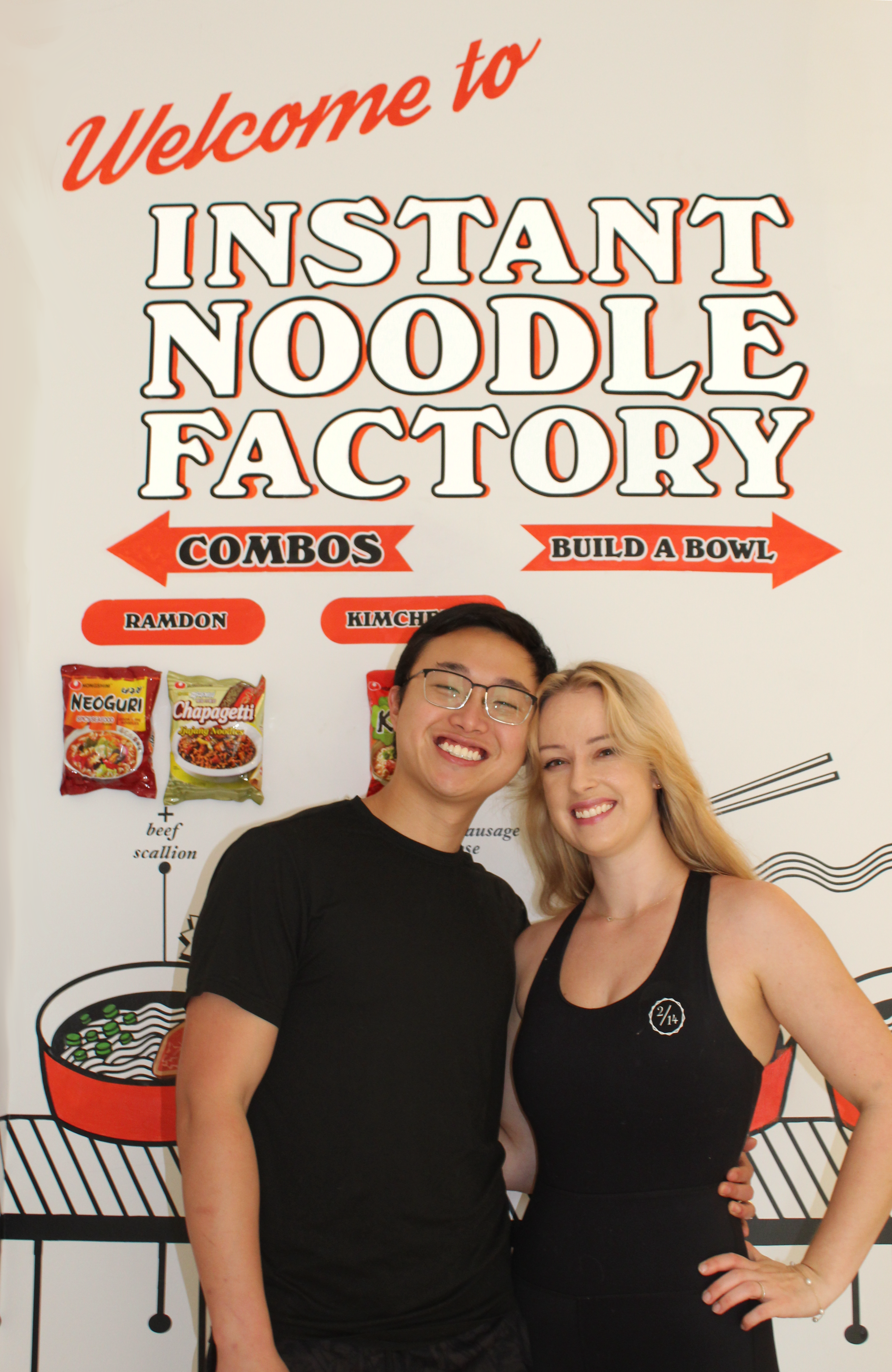 founders tat and cierra in front of the welcome to instant noodle factory sign